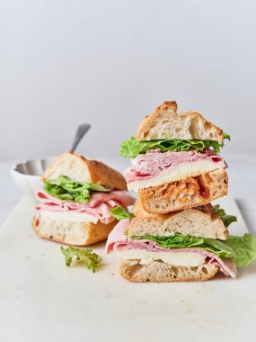 Dilimlenmiş sandviçler beyaz bir kesme tahtasına istiflenmiş. İçinde baget, jambon, mozzarella ve marul var..