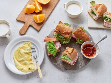 Bir öğle yemeği veya kahvaltı masasında baget, mozzarella peyniri, marul ve jambonlu dilimlenmiş sandviçler yer alır. Taze portakallar, çırpılmış yumurtalar ve bir bardak içecek mevcut..