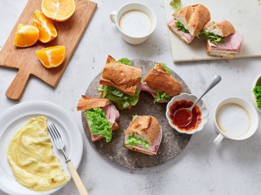 Masada baget, mozzarella, marul ve jambonla yapılan dilimlenmiş sandviçler sergileniyor. Taze portakallar ve çırpılmış yumurtalar yakınlarda, basit bir kahvaltı sahnesi yaratıyorlar..