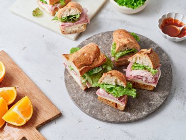 Yemek masasında baget, mozzarella peyniri, jambon, marul ve domates sosundan yapılmış sağlıklı sandviçler bulunur. Taze dilimlenmiş portakallar sahneye renk katar.