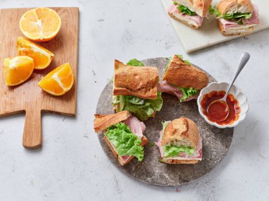 Dilimlenmiş baget sandviçler bir taş tabakta düzenlenir. Taze portakallar sandviçlerin yanında. Öğle yemeği sırasında masada küçük bir kase sos da var..