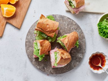 Bir yemek masasında baget, mozzarella peyniri, marul ve jambondan yapılmış sağlıklı sandviçler bulunur. Taze portakallar yakınlarda oturur, yemek düzenine renk katar..