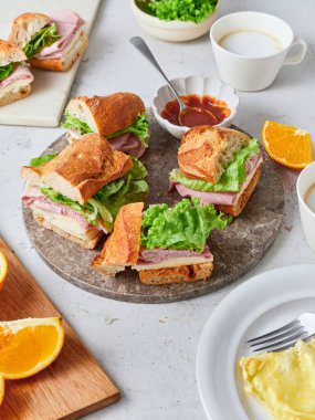 Peynirli, jambonlu ve marullu taze baget sandviçler yuvarlak tabakta. Portakal dilimleri ve çırpılmış yumurtalar masada. Kahve fincanları yakınlarda, yemek hazırlıyorlar..