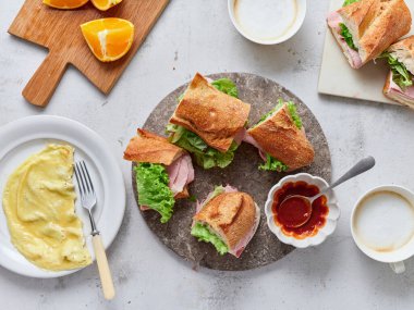 Baget, mozzarella peyniri, jambon ve maruldan yapılmış sandviçler yuvarlak bir taş tabağa konur. Yanlarında çırpılmış yumurta, dilimlenmiş portakal ve küçük bir kase domates sosu var..