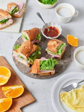 Yemek masasında baget, marul, jambon ve peynirden yapılmış dilimlenmiş sandviçler var. Taze dilimlenmiş portakal ve çırpılmış yumurta ile birlikte kahve de mevcuttur..