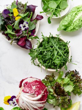 Bir mutfaktaki mermer tezgahta farklı marul türleri sıralanır. Radicchio, romaine, rogula ve diğer sebzeler mevcut. Turp, bezelye ve çiçekler ekrana eklenir..