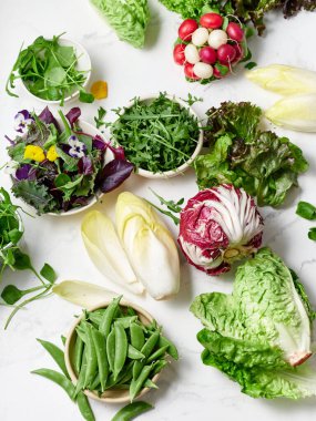 Farklı türde marul ve sebzeler bir mutfaktaki mermer tezgahın üzerinde düzenlenir. İçindekiler arasında salata yapmak için turp, bezelye ve yenilebilir çiçekler yer alıyor..