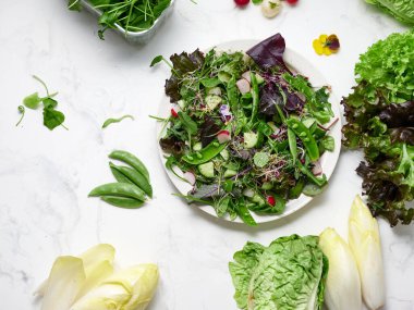 Tabakta yeşil marul, bezelye, salatalık ve turptan yapılmış taze bir salata var. Tabağın etrafındaki diğer yeşillikler mermer bir yüzeyde, basit bir vegan yemeğini gösteriyor..