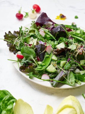 Tabakta çeşitli marullar, yeşil bezelyeler, salatalık ve turplarla taze bir bahar salatası yer alıyor. Kurgu, sağlıklı bir vejetaryen yemek seçeneğini gösteren mermer bir arkaplan içeriyor..
