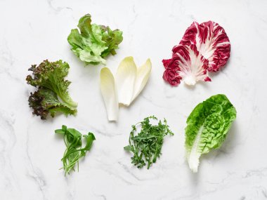 Beyaz mermer yüzeyde radicchio, romaine, rougula ve hindiba gibi farklı marul yaprakları bulunur. Yenilebilir çiçekler salata karışımına renk ve doku ekler.