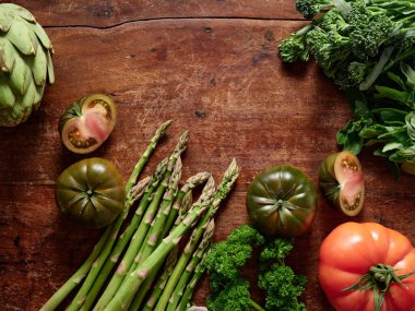 Parlak sebzeler ahşap bir yüzeyde otururlar. Sahnede domates, kuşkonmaz ve yeşillikler var. Bu ayar sağlıklı bir yemek için gerekli malzemeleri gösteriyor ve bitkilerle yemek pişirmeyi teşvik ediyor..