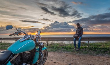 Gün batımında klasik bir motosikletin yanındaki korkuluklarda oturan bir motorcu.