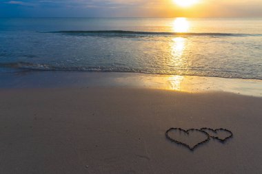 Sahilde kumlara çizilmiş iki kalp. Çerçevenin sağ tarafına yerleştirilmiş. Güneş ışığı sakin suya yansıyor ve ufuk uzaklıktan görülebiliyor. Sahne sıcak ışık, nazik dalgalar ve sakin bir atmosfere sahiptir..