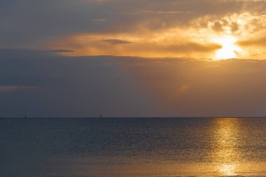 Ufuktaki yelkenli gri ve turuncu gökyüzünün altında, güneş bulutların arasında parlarken dramatik ve huzurlu bir sahne yaratıyor..
