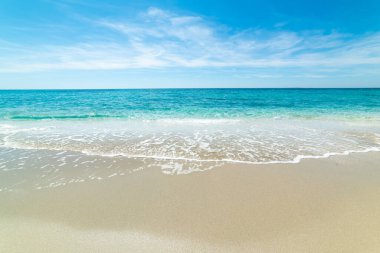 Altın kumu olan boş bir sahil ve birkaç bulutla mavi gökyüzünün altında berrak turkuaz su. Sakin denizi ve temiz ufku olan parlak kıyı manzarası.