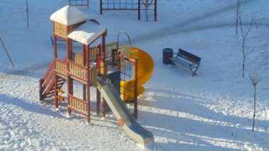 Kışın, güneşin aydınlattığı kar altında oyun parkı çocukları ve aileleri bekliyor, zaman akıp gidiyor.