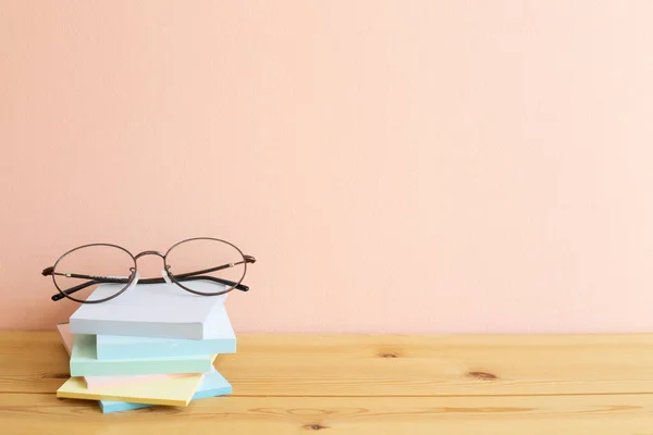 Glasses and stack of colorful memo pad, sticky notes on wooden desk. Pink background, copy space. Work and study place