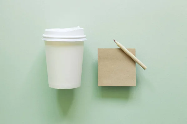 Brown kraft memo pad, pencil, paper coffee cup on green background. top view, copy space