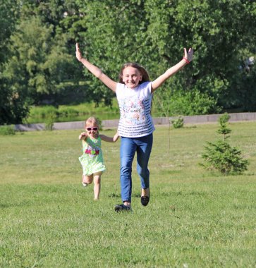 Çocuklar yeşil çimlerde zıplayarak eğleniyorlar. İki kız çimlerde koşuyor. Mutlu aile takımı. Kız kardeşler yaz tatillerinin tadını çıkarırlar. Çocukların keyfi yerinde.