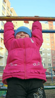 Yatay çubukta asılı duran kız. Tuhaf çocukluk. Çocuk yatay çubukla oynuyor. Oynarken çocukça bir yüz ifadesi. Yatay çubukta asılı duran çocuk gülümsüyor