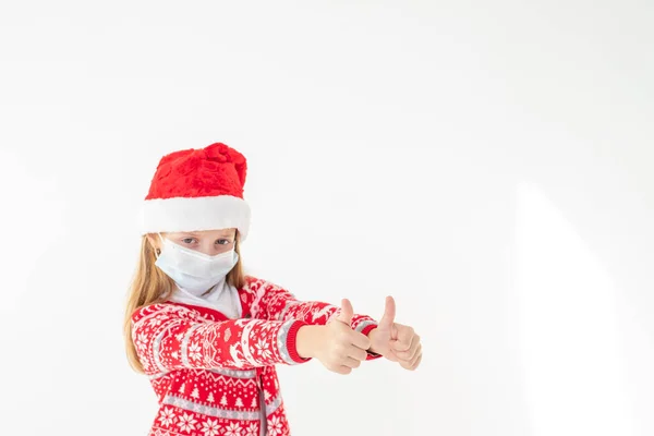 Retrato de una hermosa niña con sombrero de Santa sonriendo y mostrando los pulgares hacia arriba, aislado sobre fondo blanco con espacio para copiar. celebrando feliz Año Nuevo. Seguridad infantil distancia social —  Fotos de Stock