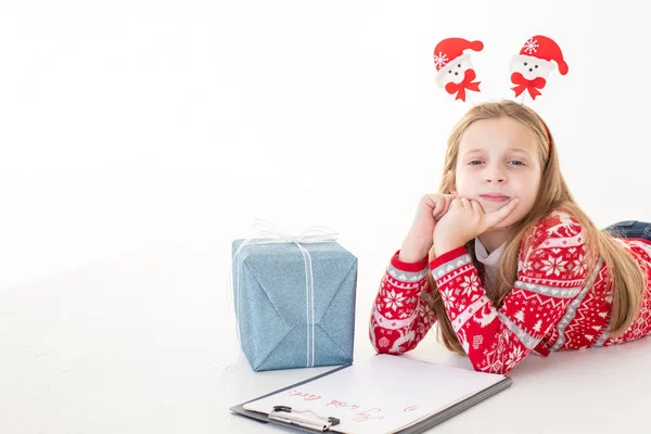 생각깊은 어린 소녀는 하얀 배경에 산타클로스에게 편지를 쓴다. 산타를 위한 특별 명령이야. 선물 옆에 누워 있는 소녀 — 스톡 사진