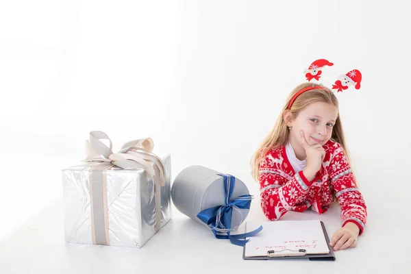 Niña reflexiva escribe carta a Santa Claus sobre fondo blanco.Metas planes sueños hacen que hacer lista para el concepto de Navidad de año nuevo. Órdenes especiales para Santa. chica acostada cerca de los regalos —  Fotos de Stock