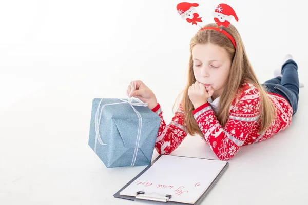 Kära tomte brev skrivet av ett barn för julen. Koncentrerad unge är inblandad i processen att tänka över alla hennes önskningar och önskningar. Pretty Girl hålla urklipp och papper nära presentförpackning. — Stockfoto