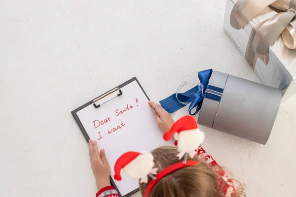 Querida carta de santa escrita por un niño para Navidad. niño concentrado está involucrado en el proceso de pensar en todos sus deseos y deseos. Pretty Girl sujetar portapapeles y papel cerca de la caja de regalo. —  Fotos de Stock