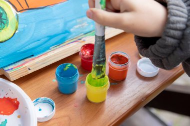 Çocuk resim malzemeleriyle masayı boyuyor. Küçük çocuğu ve yaratıcılık masasını boyamak. Çocuklar yaratıcılığı etkiler. Evde eğitim konsepti çizimi. Kavanozlar boya, boya fırçaları, paletler..