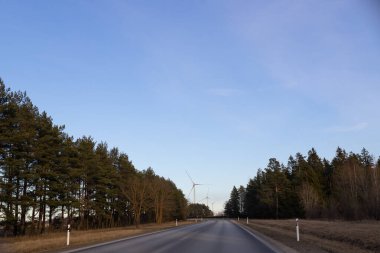 Yol orman ve yel değirmenlerinin çok ötesine gidiyor. Rüzgar türbini. Önde boş bir yol, bulutlu mavi bir gökyüzü. Alternatif enerji kaynağı, üretim ve güç üretimi. Ekoloji ve özgürlük kavramı.
