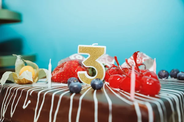 Birthday-anniversary cake with gold candle showing number 3.Chokolate cake with fruits and a candle with the number three on blue background. selective focus. — ストック写真