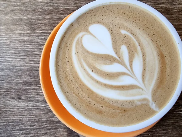 Kopi dihiasi dengan hati dan daun, selamat pagi. Hot Cappuccino dengan cangkir oranye di atas meja kayu di warung kopi. Salin ruang — Stok Foto