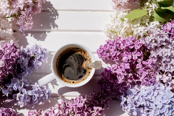 黒コーヒーの朝のカップと白い背景に緑の葉で紫色のライラックを開花の枝。コーヒーの白いカップ、ライラックの美しい花束と組成。春と快適さの概念. — ストック写真