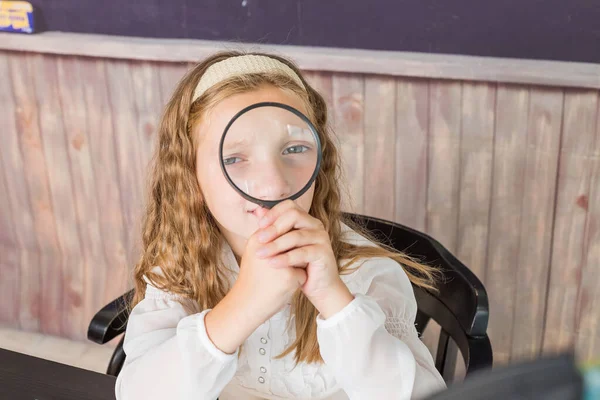 Education and school concept - little girl with magnifier at school. schoolgirl sitting in the classroom. Child in an elementary school. Education and learning for kids. — Stock Photo, Image