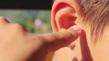 Genç bir çocuk işaret parmağıyla kulağının içini dikkatlice temizliyor, pencerenin yanında doğal aydınlatmayla otururken kişisel hijyene odaklanıyor.