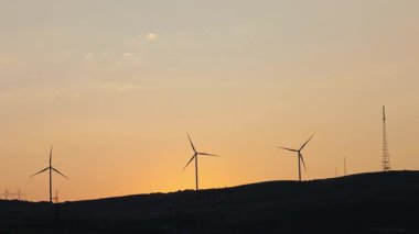 Siluetli rüzgar türbinleri canlı günbatımı gökyüzüne karşı dönüyor. Manzaralı yamaçlarda temiz yenilenebilir enerji üretiyorlar.