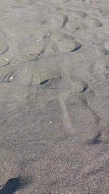 Yazın çarpıcı bir volkanik plajın nemli, abanoz gibi kıyı şeridinde çıplak ayakla yürüyen bir figürün perspektifi kumlara taze ayak izleri bırakıyor.