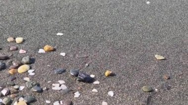 Yaklaşan okyanus gelgiti yavaş yavaş küçük çakıl taşları ve deniz kabuklarıyla süslenmiş gölgeli bir volkanik kumsalı yutuyor. Yakından bakınca, yumuşak dalgaların kıyıya vuruşunu yakalıyor.