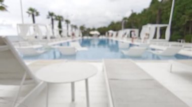 Prestijli bir tatil köyünde üst düzey bir yüzme havuzunun bulanık görüntüsü. Havuzun kenarında zarif beyaz sandalyeler ve gölgeli kabinler var. Hepsi bulutlu bir gökyüzüne karşı dizilmiş.