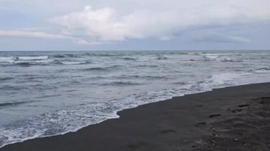 Sakin okyanus dalgalarını içeren bulutlu deniz manzarası yavaşça siyah, volkanik bir kumsal şeridine vuruyor. Melankolik ve izole kıyı manzaraları