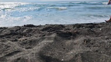 Genç kadın bacakları, siyah bir kumsalın kıyılarında yalın ayak yürüyor. Işıldayan okyanus dalgaları arka planda görünür.