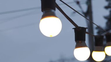 Warm light bulbs on a string glowing brightly. Outdoor decoration for a party or event against a moody, blurred winter background