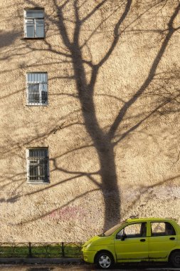 Duvarında bir ağacın gölgesi olan şehir manzarası. Büyük bir evin arka planında küçük sarı bir araba.