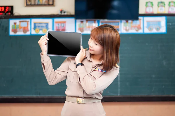 female thai teacher in government uniform standing in front of classroom holding ipad