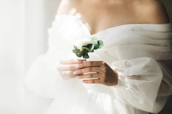 Close-up das mãos noivas mantém um boutonniere. Mulher com um lindo vestido branco. Preparativos para noivas. Conceito da manhã de casamento. Ênfase nas mãos e decoração. — Fotografia de Stock