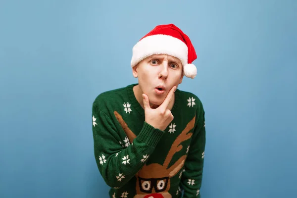 Amazed guy in Christmas clothes stands on a blue background and looks into the camera with a thoughtful shocked face. Young man in santa hat looks into camera and is surprised.