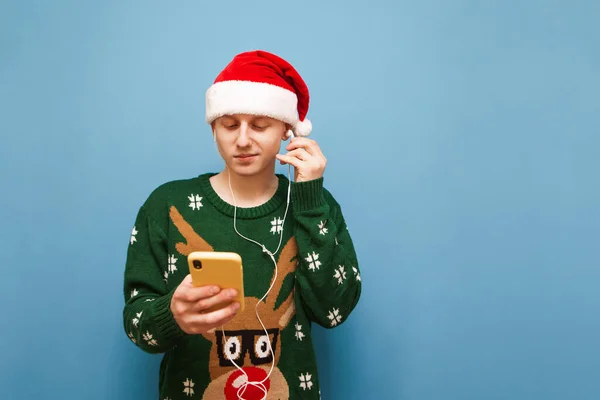 Handsome young man in sweater with deer and santa hat stands on blue background with smartphone in hand and listens to music in headphones. Guy in a sweater and a santa hat listens to Christmas music