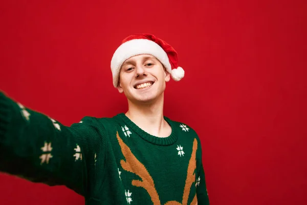 크리스마스 배경으로 셀카를 만들고 카메라를 들여다보며 젊은이 스웨터입고 모자쓴 긍정적 — 스톡 사진