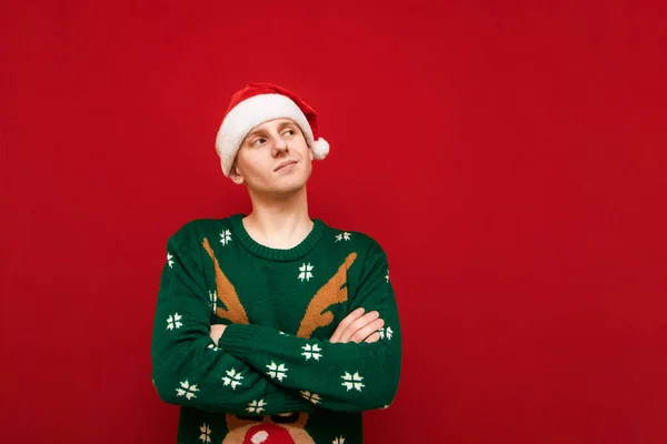 Cute young man in santa hat and green christmas sweater stands on red background, looks away at blank space and smiles. Positive guy in christmas clothes isolated on red background.